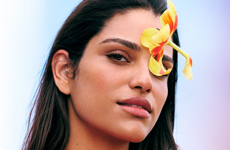 Foto con fondo celeste. Primer plano del lateral del rostro de una mujer, cabello oscuro, oreja con piercing y una flor naranja detrás de la oreja.